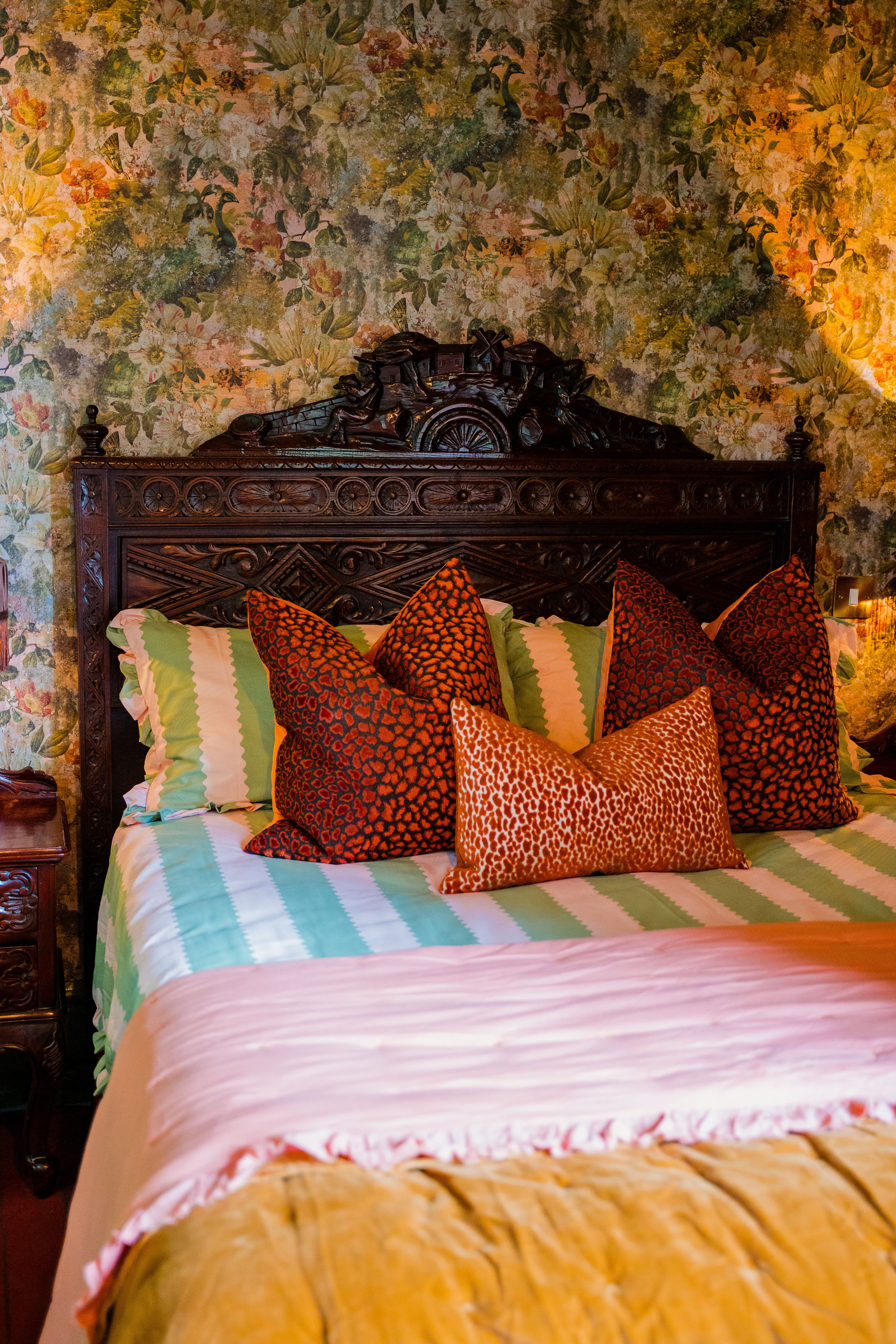 Close up of wooden bed featuring maximalist bedding against a patterned wallpaper.